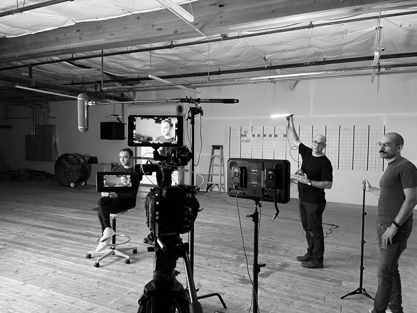 A film crew prepares to record a seated man in a large, industrial-style studio, with a camera operator adjusting a small light while another crew member stands nearby holding a stand.