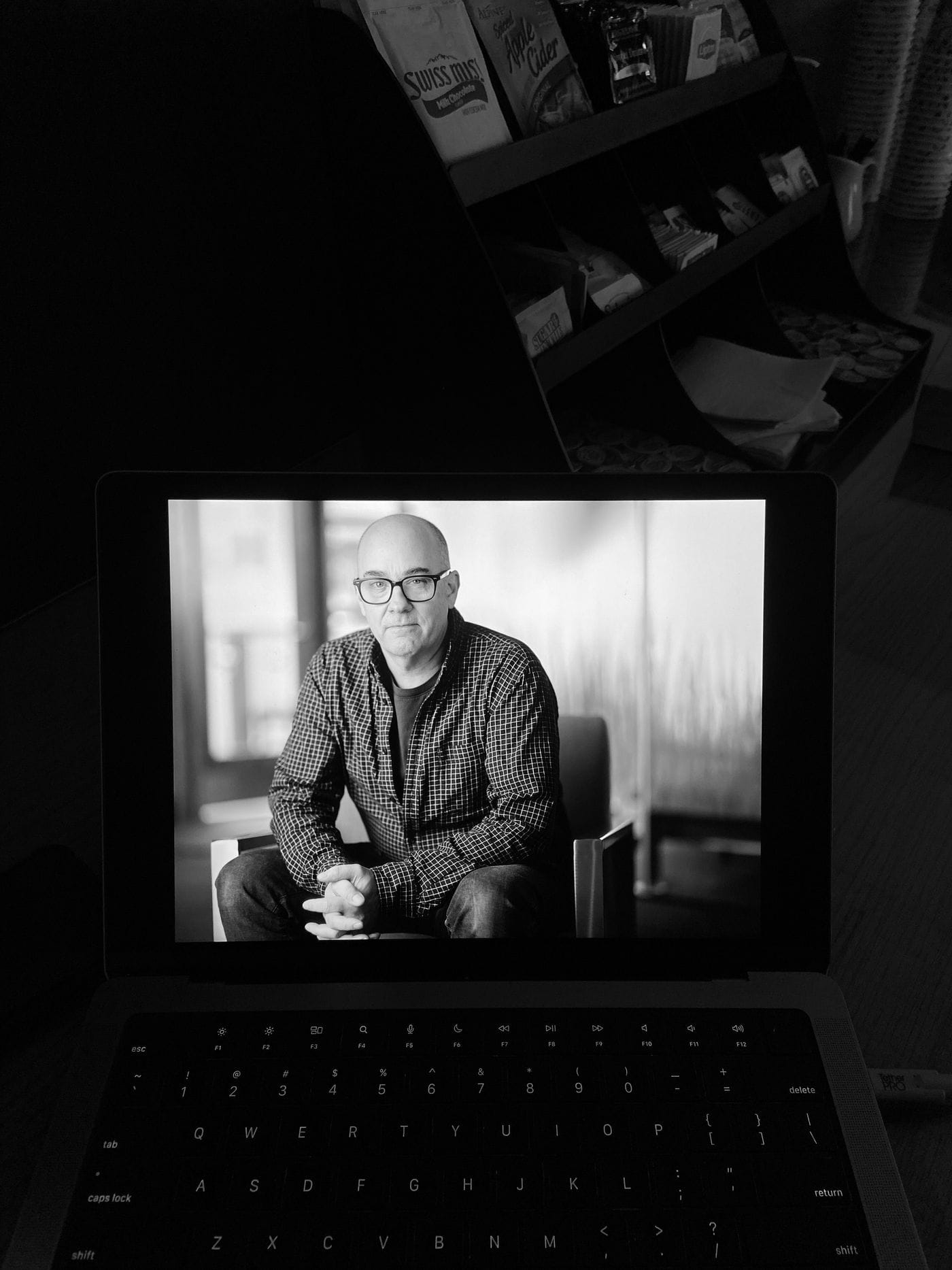 A black and white portrait of a bald man with glasses, wearing a plaid button-down shirt and sitting with his hands clasped, is displayed on a laptop screen placed in front of a shelf stocked with coffee and tea packets.