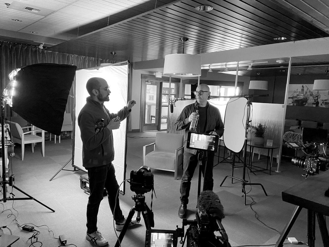 A filmmaker gestures while speaking to a man holding a water bottle in an indoor studio space set up with professional cameras, lighting equipment, and a large white backdrop.