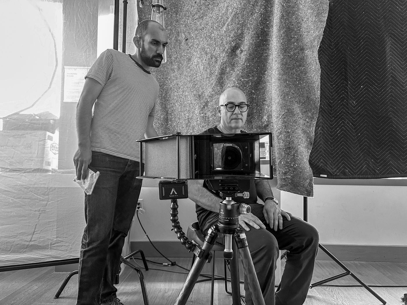 A bald man wearing glasses sits on a chair in front of a camera mounted on a tripod while a man in a striped shirt stands beside him in a studio setting.