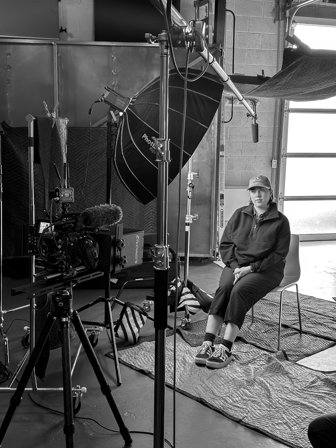 A person wearing a John Deere baseball cap and a dark quarter-zip sweater sits on a metal chair in a studio, facing a professional video camera setup equipped with a shotgun microphone and a large softbox light.