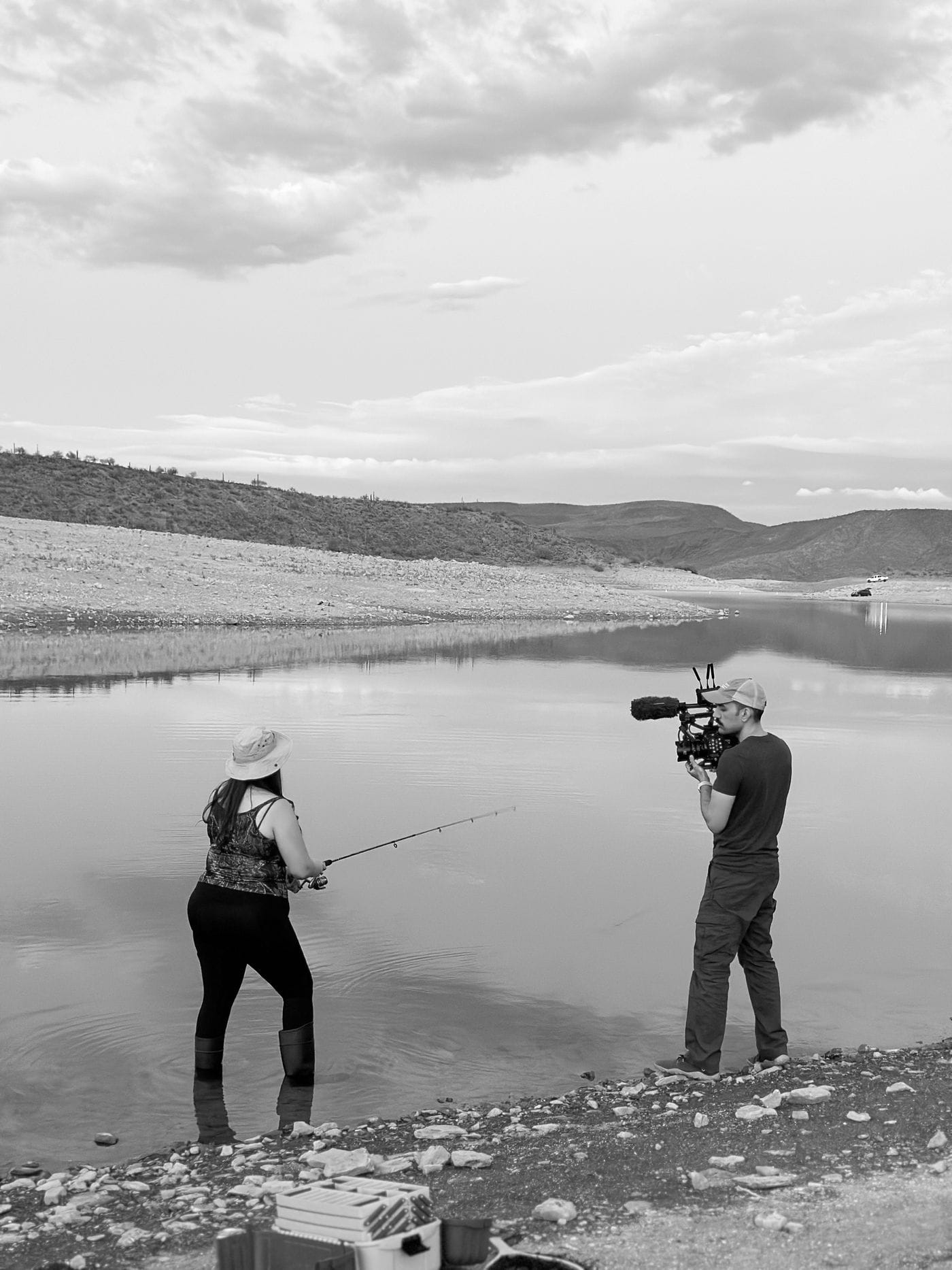 A videographer films a woman wearing a sun hat and waders as she fishes in a calm, scenic lake surrounded by hills under a cloudy sky.