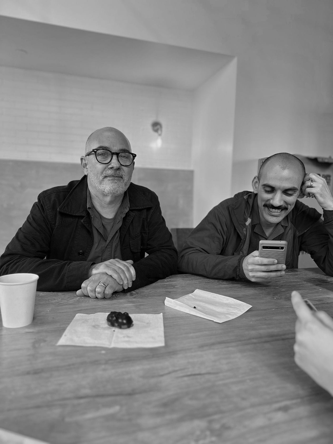 Two men sit at a wooden table in a cafe, with one looking at the camera while the other smiles at a smartphone, accompanied by coffee cups and pastries on napkins.