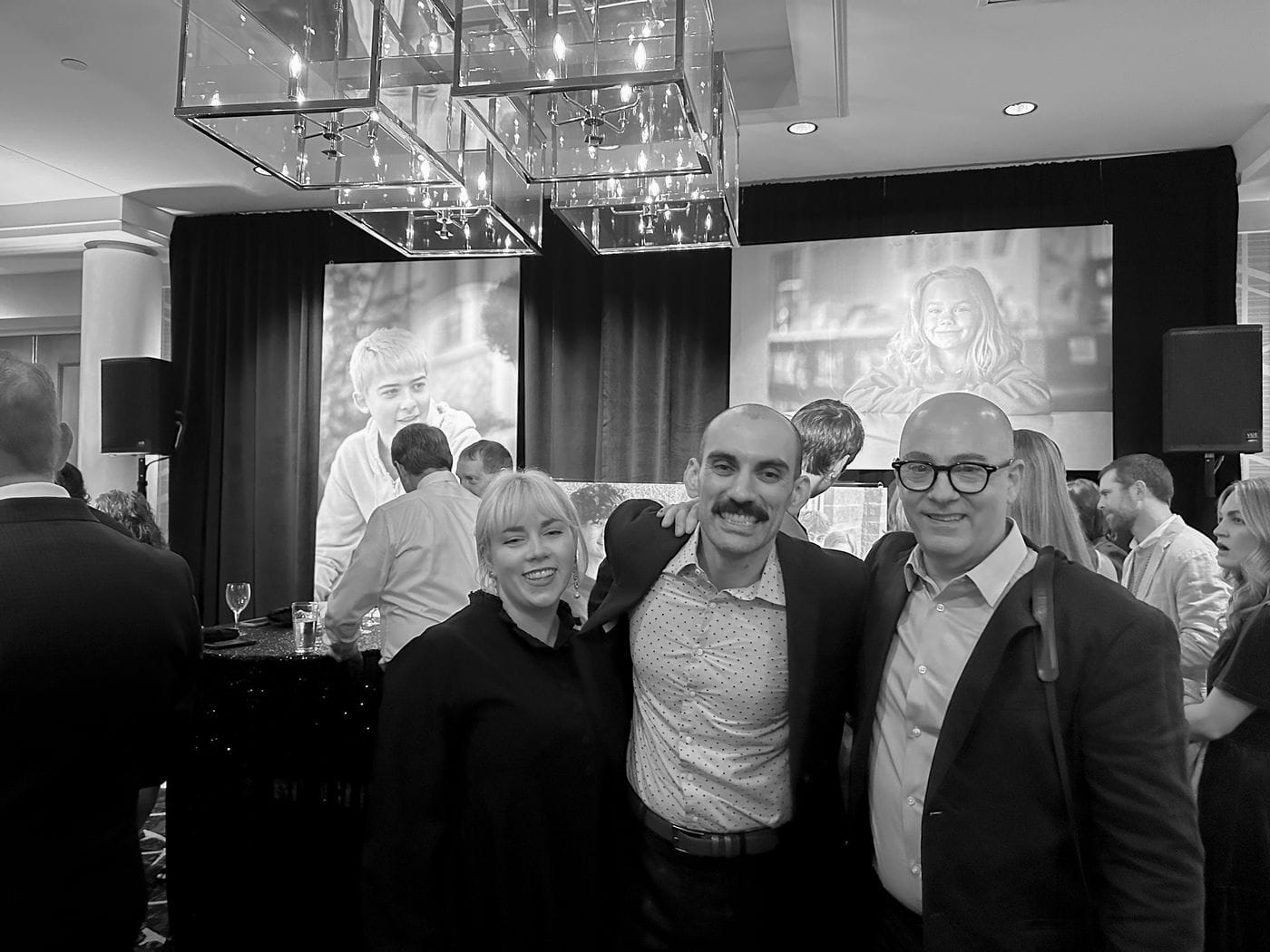 A woman and two men pose for a photo at an indoor event, standing in front of large black-and-white portraits and a decorative light fixture.