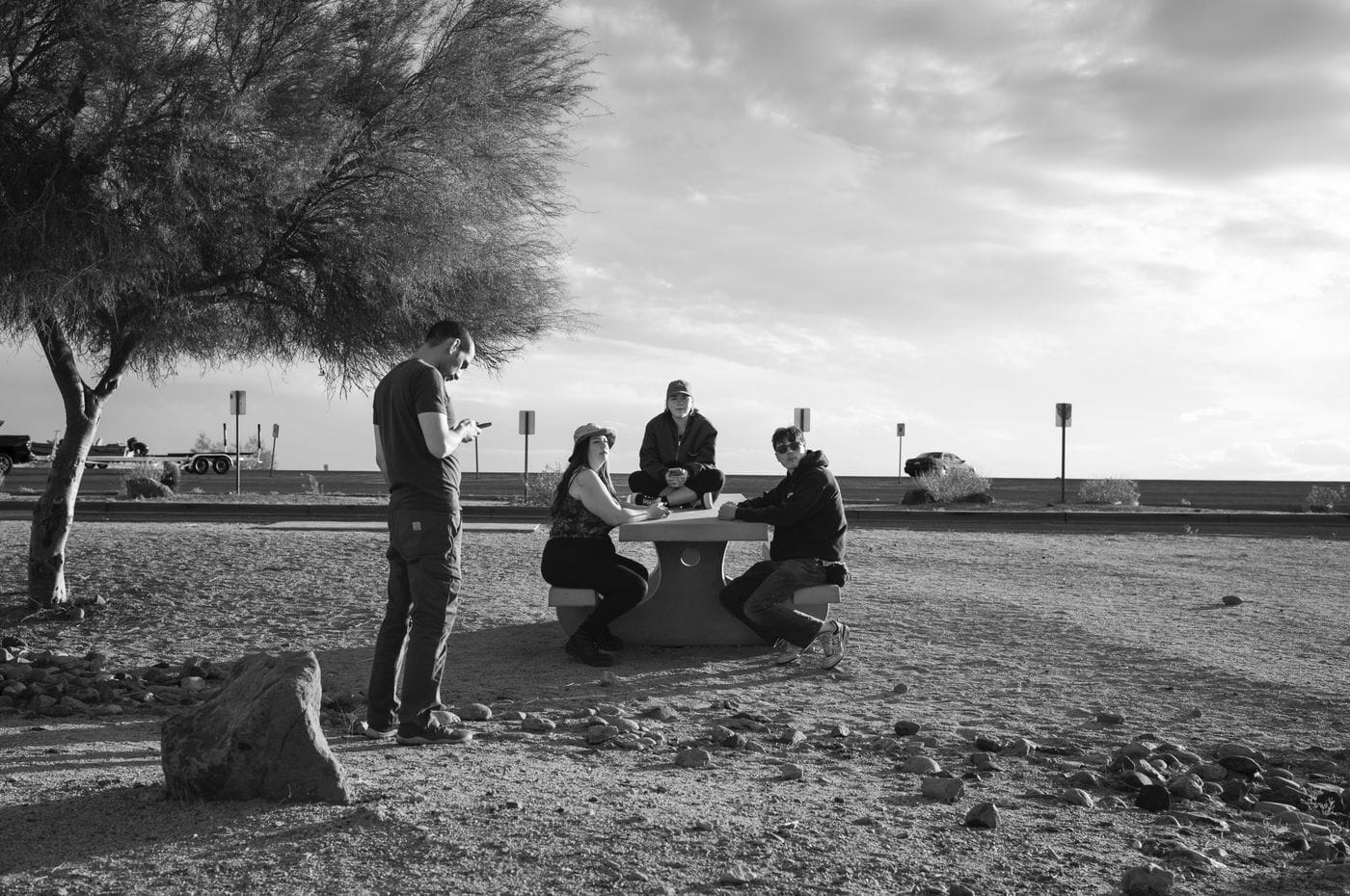 A group of four people gather at a concrete picnic table in a sun-drenched, gravel-covered outdoor area under a large, bare tree while a man stands nearby looking at his phone.