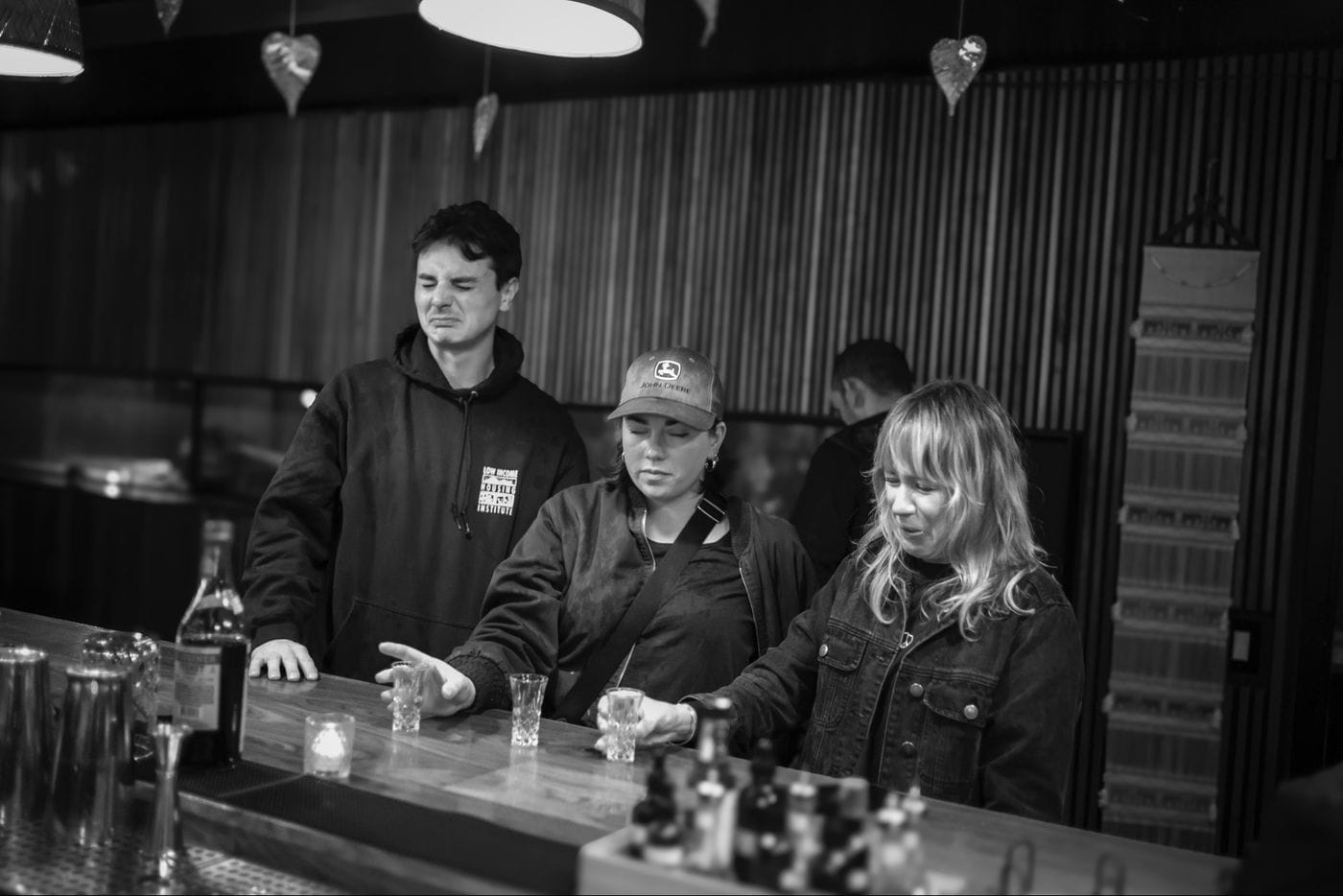 Three people stand at a bar, making grimacing expressions while reaching for small shot glasses.