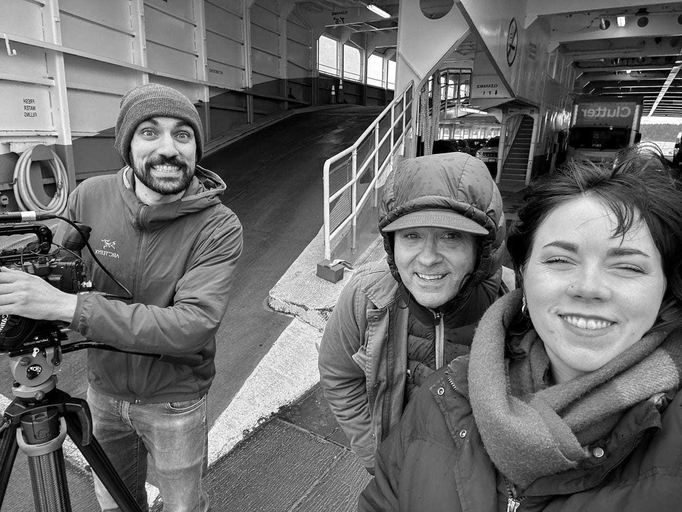 A black-and-white selfie of three people on a ferry car deck, with a man on the left holding a professional video camera on a tripod, a man in a hooded jacket in the center, and a smiling woman on the right.