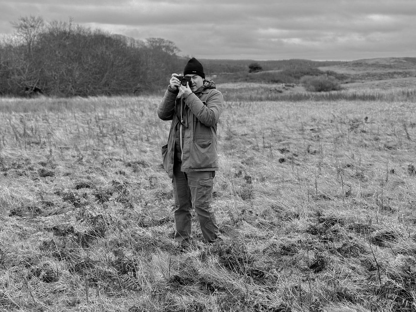 A man wearing a beanie and a jacket stands in a vast, grassy field under a cloudy sky, holding a camera up to his face to take a photograph.