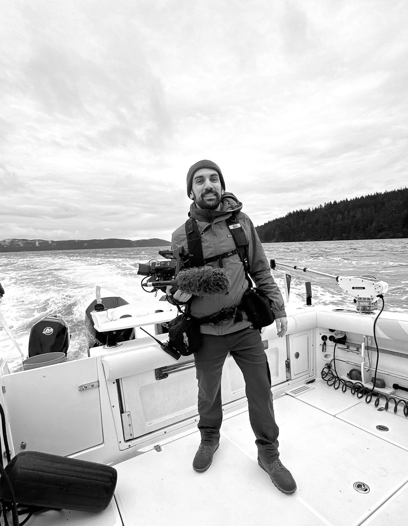 A smiling man in a beanie and jacket stands on a boat deck, holding a professional camera with a shotgun microphone, with water and a distant forested shoreline in the background.