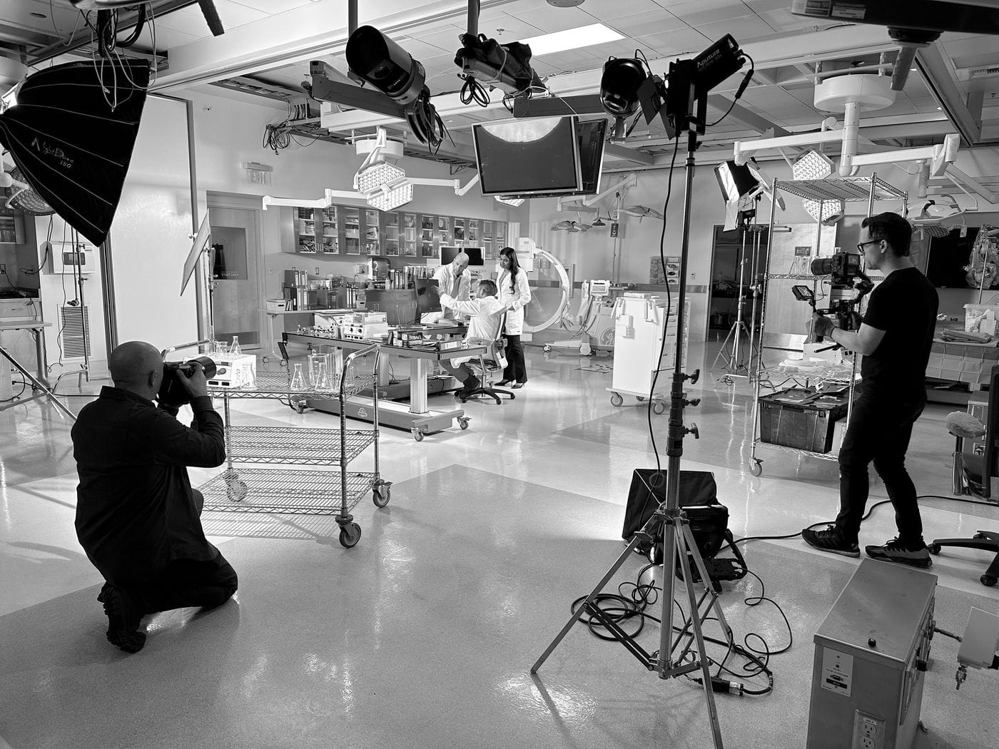 A film crew captures a scene in a sterile, clinical setting with three people in lab coats gathered around a workstation as a cameraman films them and a photographer kneels to take stills.