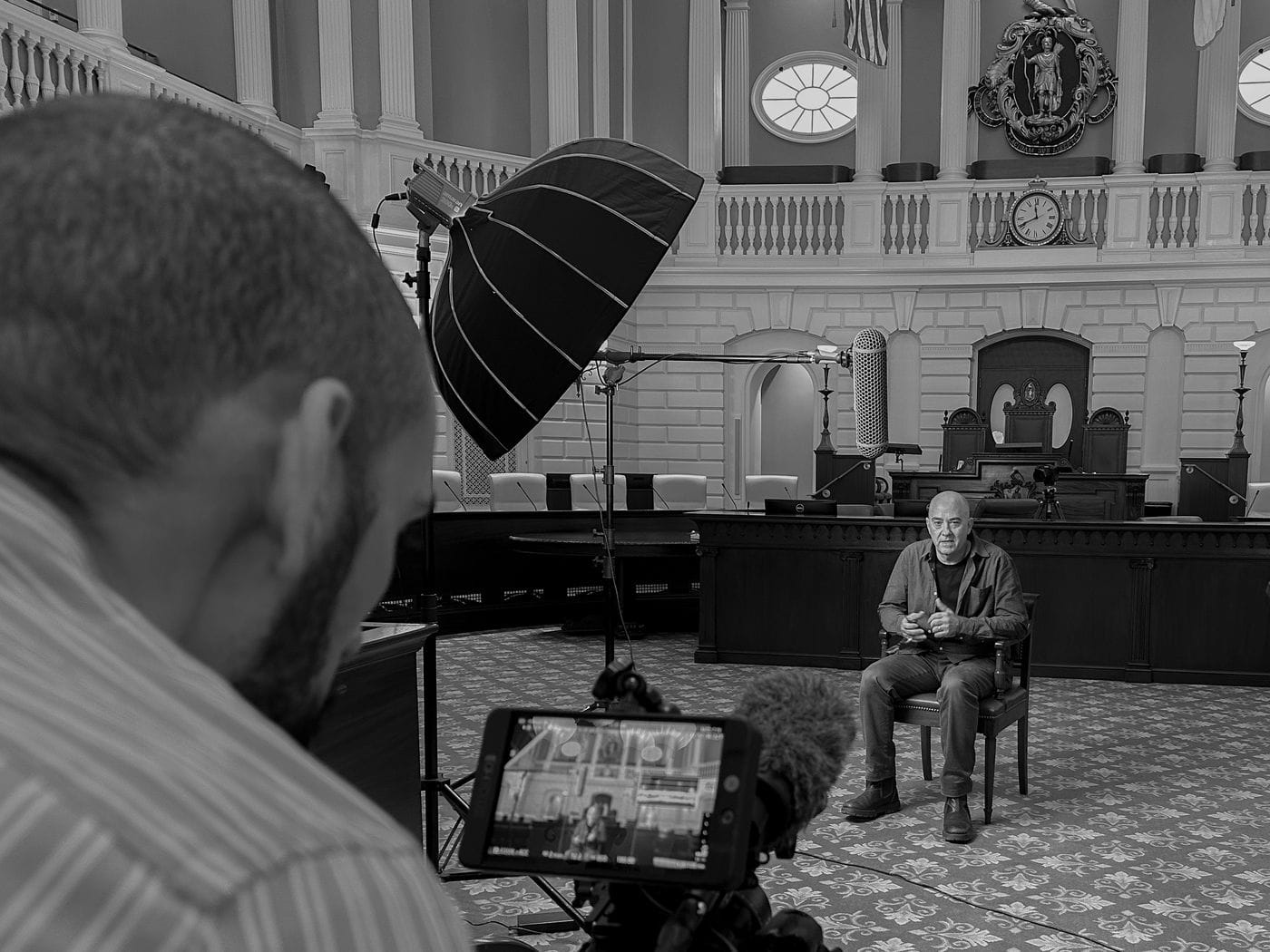 A videographer in the foreground films a bald man sitting in a chair for an interview inside a grand, historic chamber with decorative moldings, a coat of arms, and a large analog clock on the wall.