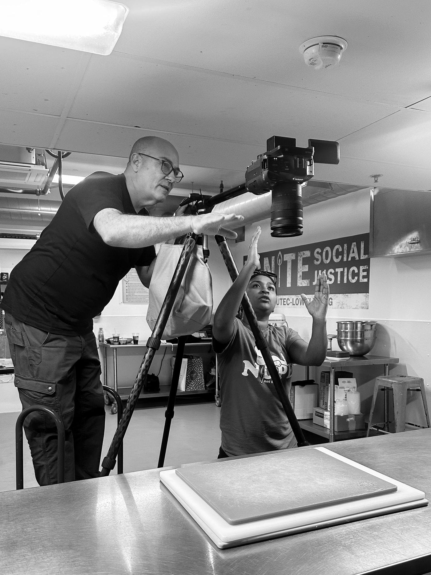 A man with glasses stands behind a tripod-mounted camera, gesturing to a woman who stands in front of it in a kitchen setting, as they coordinate a shot above a cutting board.