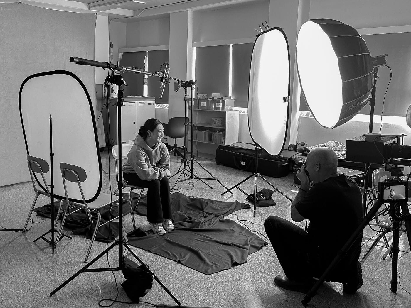 A bald photographer in a black shirt kneels to photograph a smiling woman wearing a hoodie who sits in a chair in a studio lit by large softboxes and a boom mic overhead.