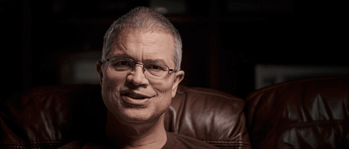A middle-aged man with short, graying hair and glasses smiles while sitting on a brown leather couch in a dimly lit room.
