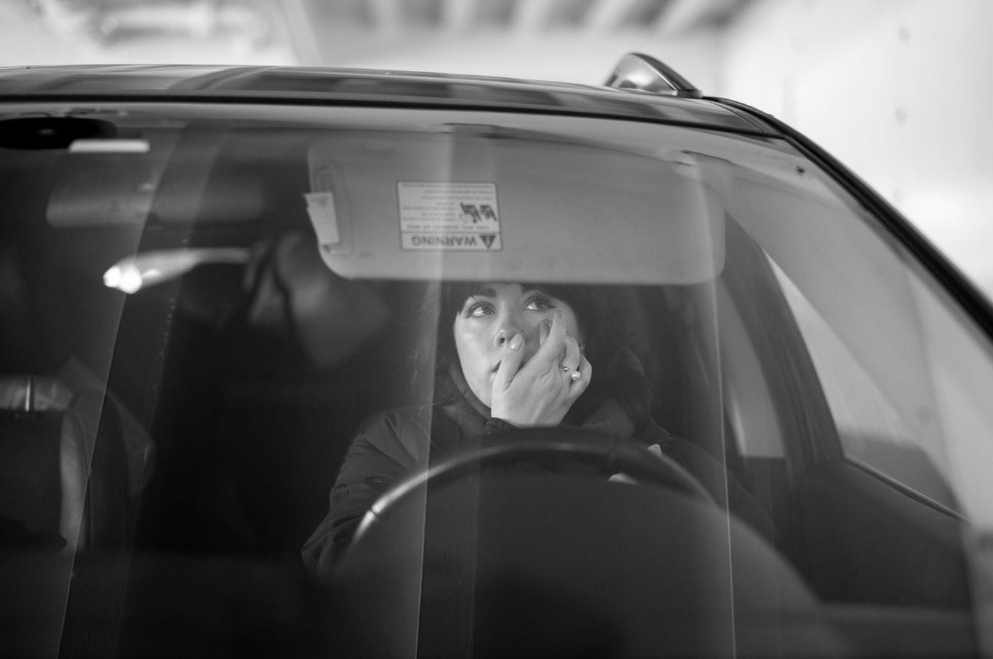 A young woman sits in the driver's seat of a car, looking up while adjusting a contact lens in her eye.