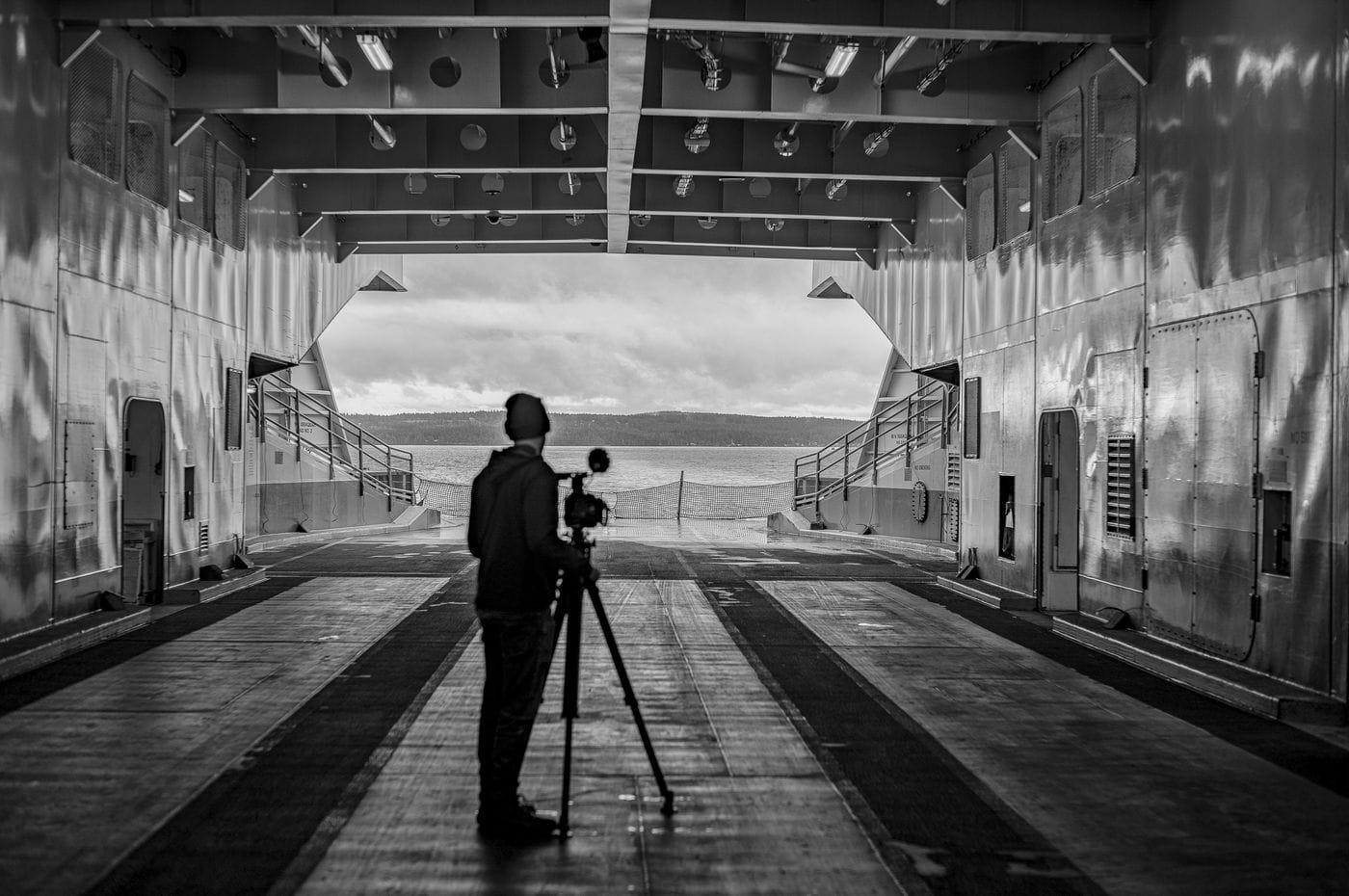 A person in a beanie and jacket stands on a ferry car deck behind a camera mounted on a tripod, looking out toward a body of water and distant shoreline under a cloudy sky.