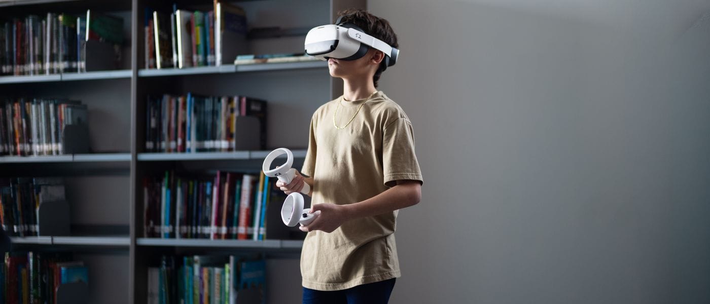 A young boy wearing a VR headset and holding controllers stands in front of a bookshelf in a library.