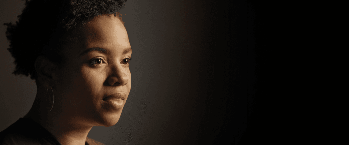 A side-profile close-up of a Black woman with short, curly hair looking off-camera against a dark, moody background with subtle directional lighting.
