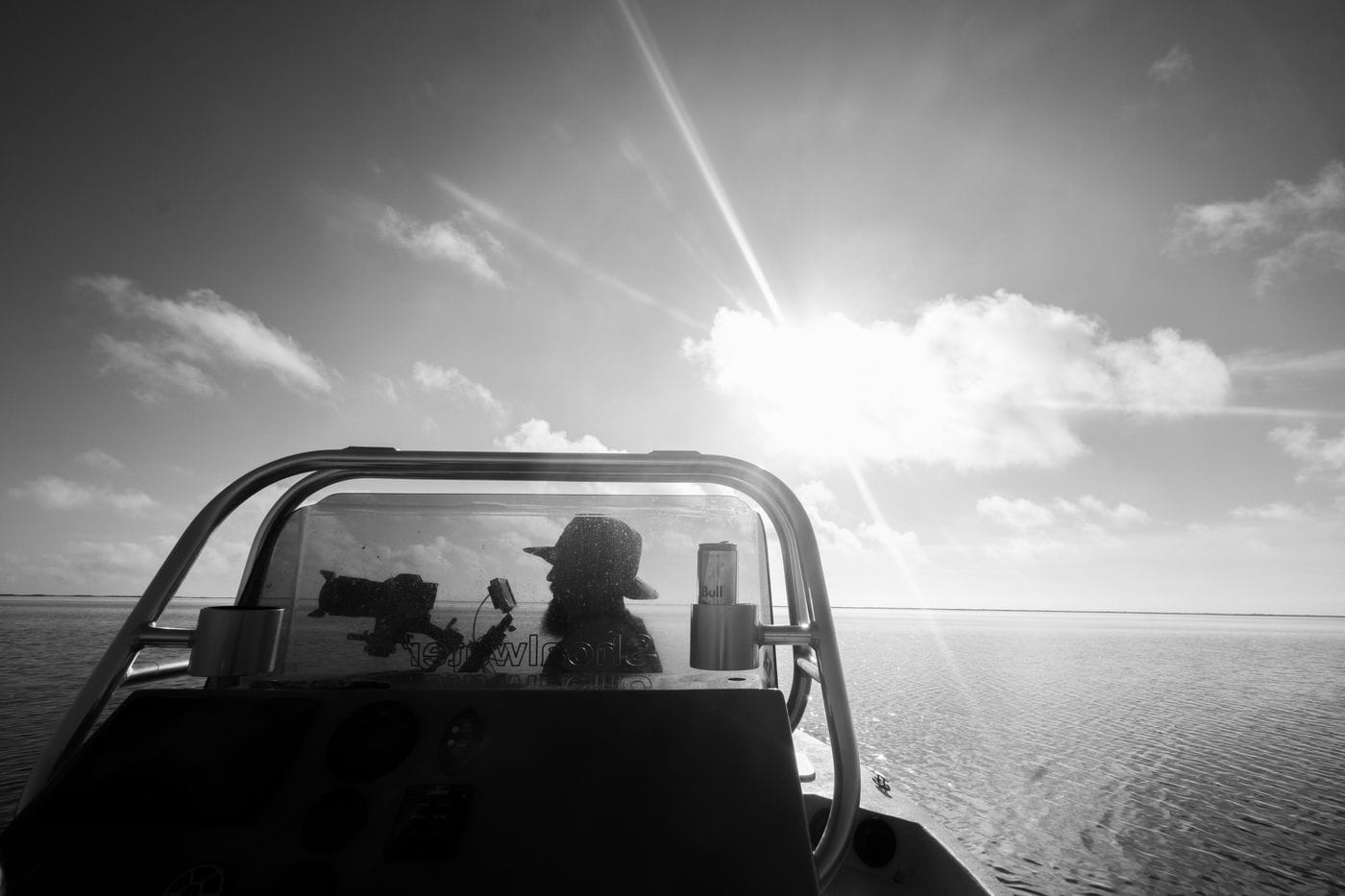 A person wearing a wide-brimmed hat sits behind a boat's windshield, silhouetted against a bright, sunlit sky over a calm expanse of water.