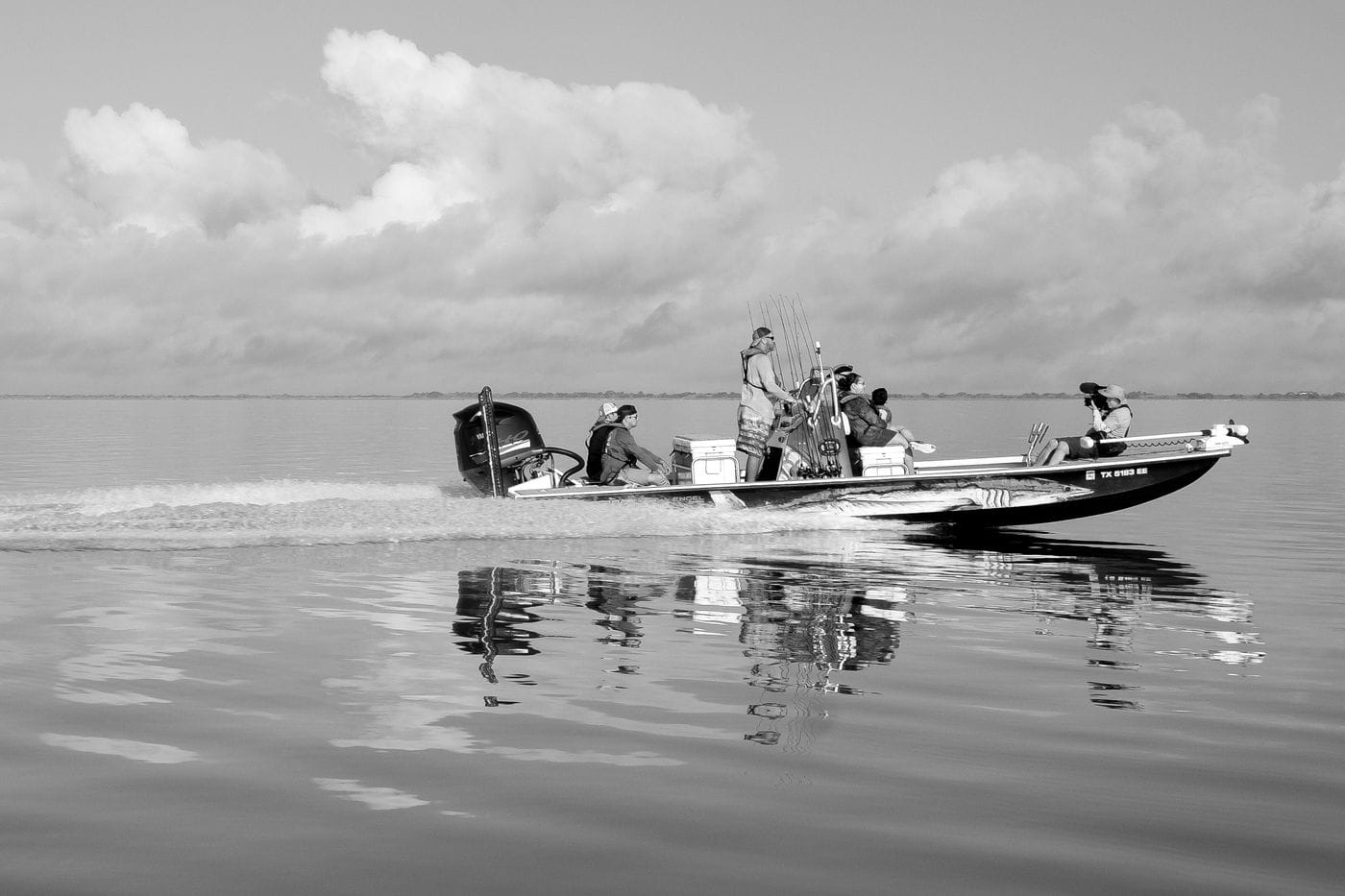A group of people, including one filming with a camera, ride in a motorboat across a calm, reflective body of water under a cloudy sky.