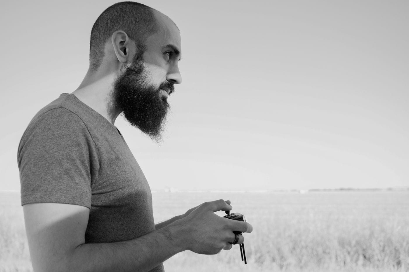 A side profile of a man with a thick beard wearing a t-shirt as he stands in a field holding a drone remote control.