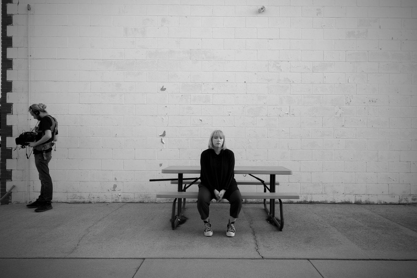 A blonde woman wearing a black long-sleeved shirt and dark pants sits centered on a picnic bench against a white brick wall, while a man with a ponytail and headphones stands to the left operating a large video camera.