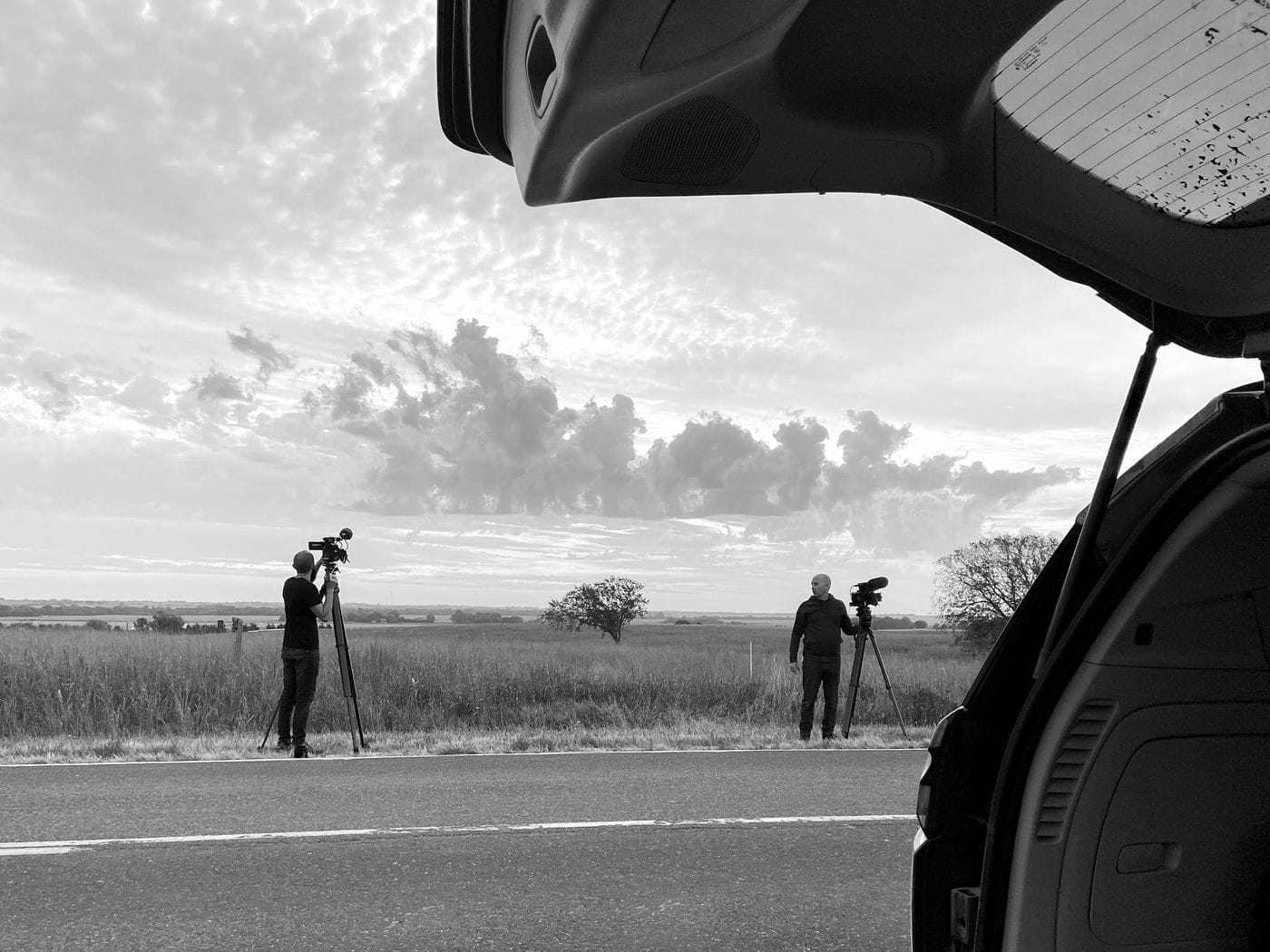 Two cameramen with tripods stand on the shoulder of a country road during sunset, as viewed from the open trunk of a vehicle.