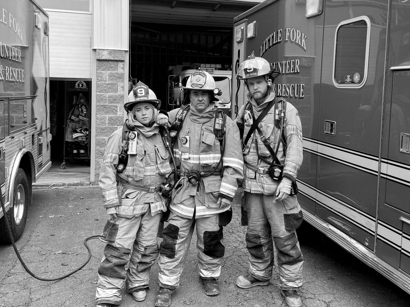 Three firefighters in full protective gear and helmets stand in front of a fire station between two emergency vehicles.