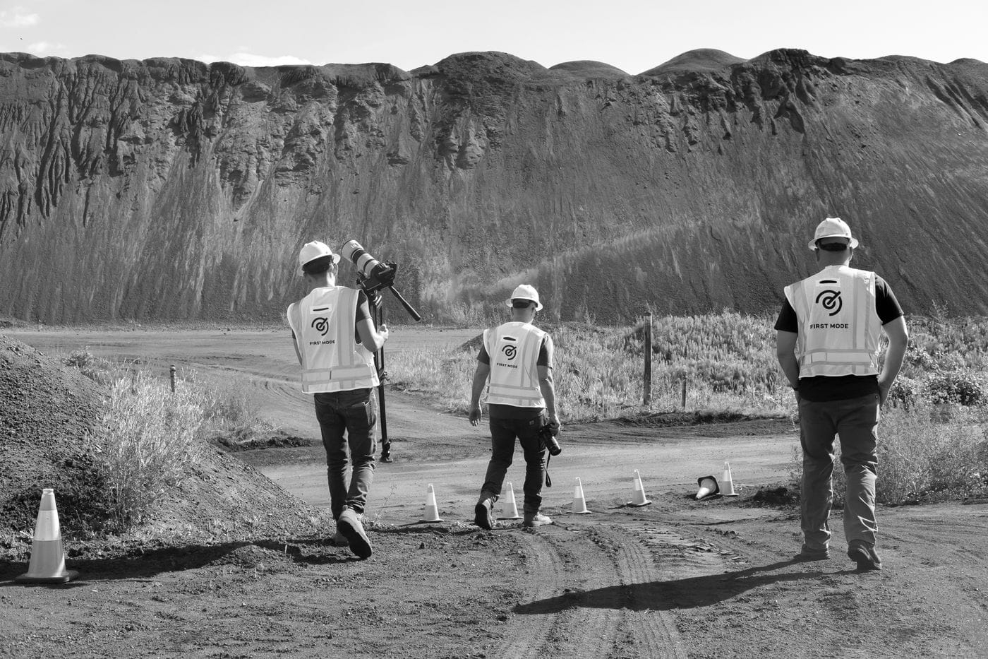Three people wearing hard hats and branded First Mode safety vests walk along a dirt path toward a large, craggy mountain of dark rock, with the person on the left carrying a tripod-mounted professional camera.