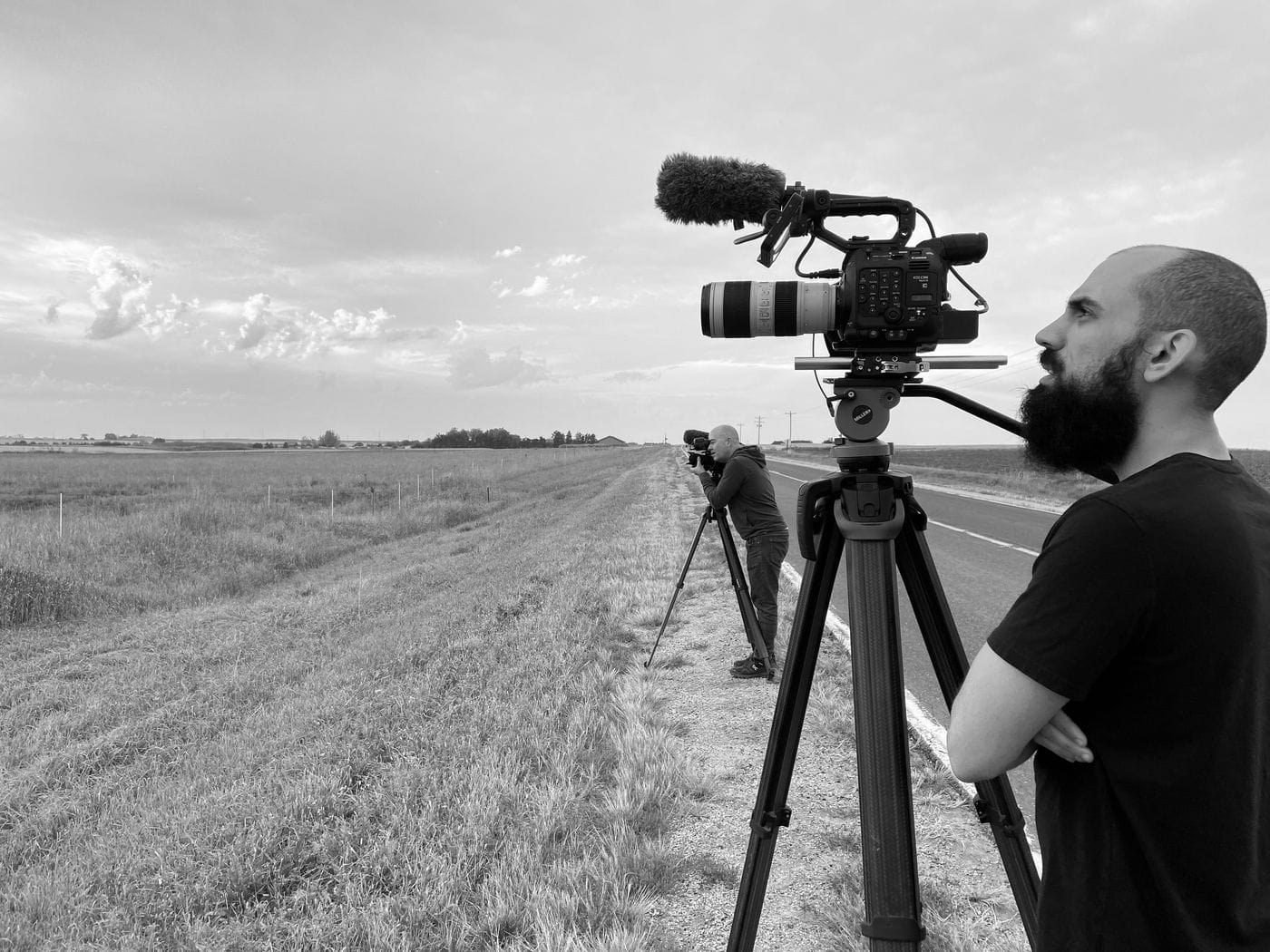 Two men with beards stand along the shoulder of a rural road, each filming the horizon with a professional video camera mounted on a tripod.