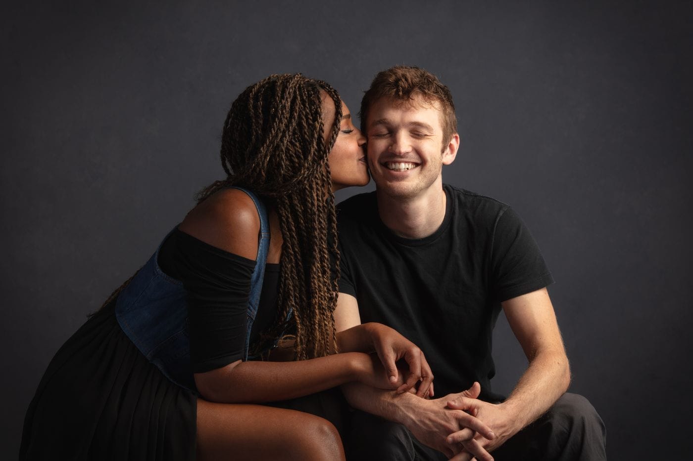 A woman with braided hair kisses a smiling man on the cheek while they sit together holding hands against a dark background.