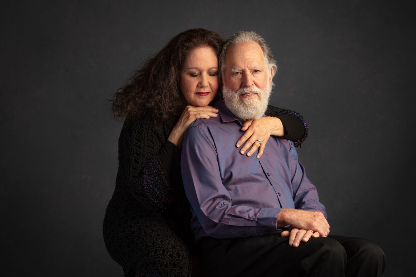 A woman with long, dark, wavy hair, wearing a black textured top, embraces a bearded man in a purple button-down shirt from behind as they pose together against a dark gray background.