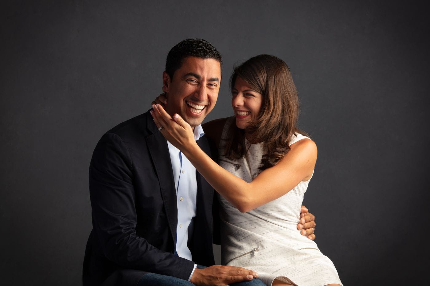 A man in a navy blazer and a woman in a white sleeveless dress laugh together while seated against a dark gray background, with the woman gently touching the side of the man's face.