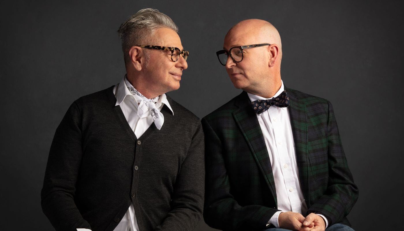 Two men with glasses face each other against a dark gray background, one wearing a cardigan with a neckerchief and the other wearing a plaid blazer with a bow tie.