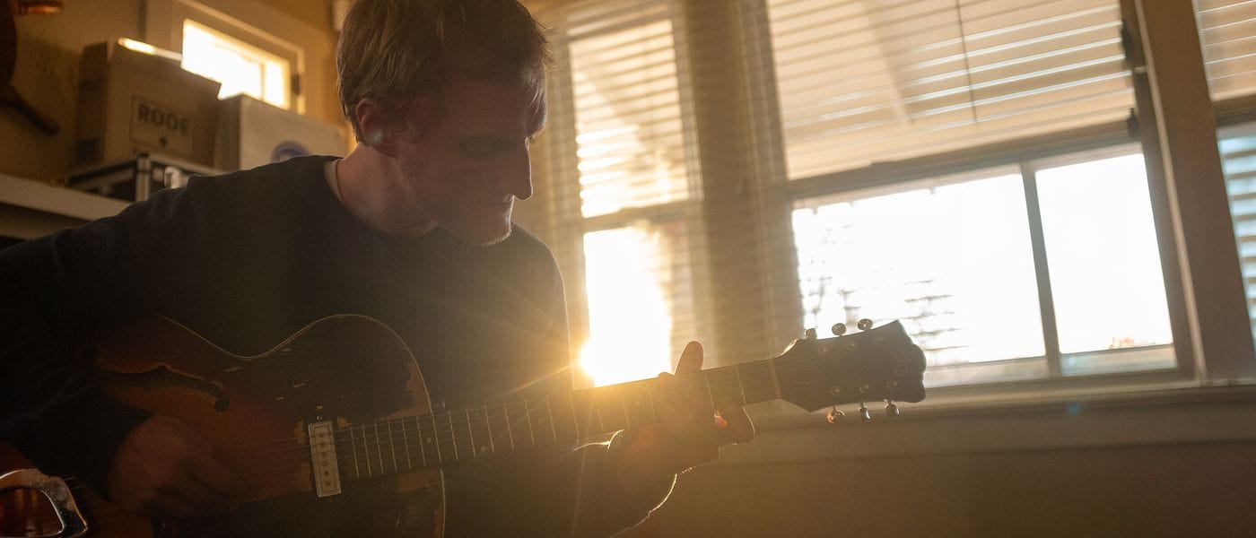 A person wearing a dark sweater plays an acoustic electric guitar in a room filled with golden backlight streaming through large windows.
