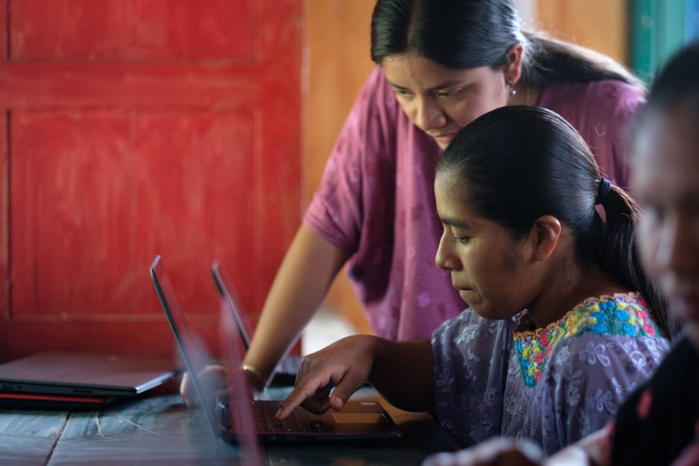 A woman leans over a seated indigenous woman to assist her with typing on a laptop in a brightly lit indoor space with red wooden walls.