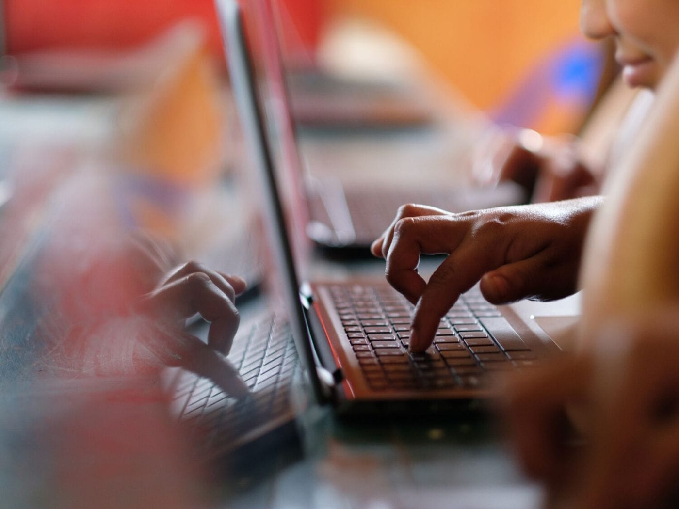 A close-up, shallow-depth-of-field shot captures a person's hand typing on a laptop keyboard, with a clear reflection of the hand and keyboard visible on the reflective surface of the screen.