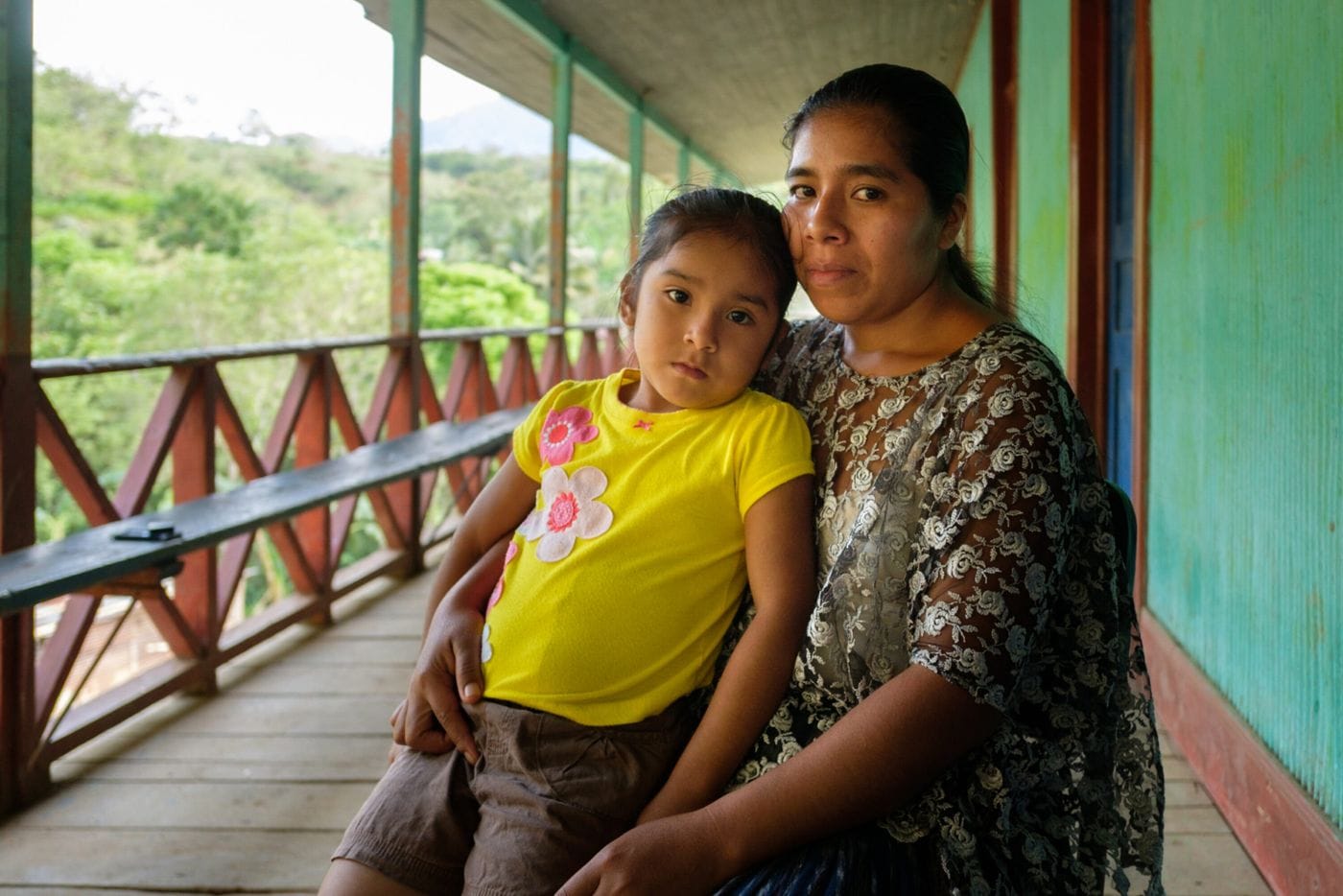 A woman with dark hair and a lace top sits on a wooden porch, holding a young girl in a yellow flower-decorated shirt on her lap as they both look toward the camera.