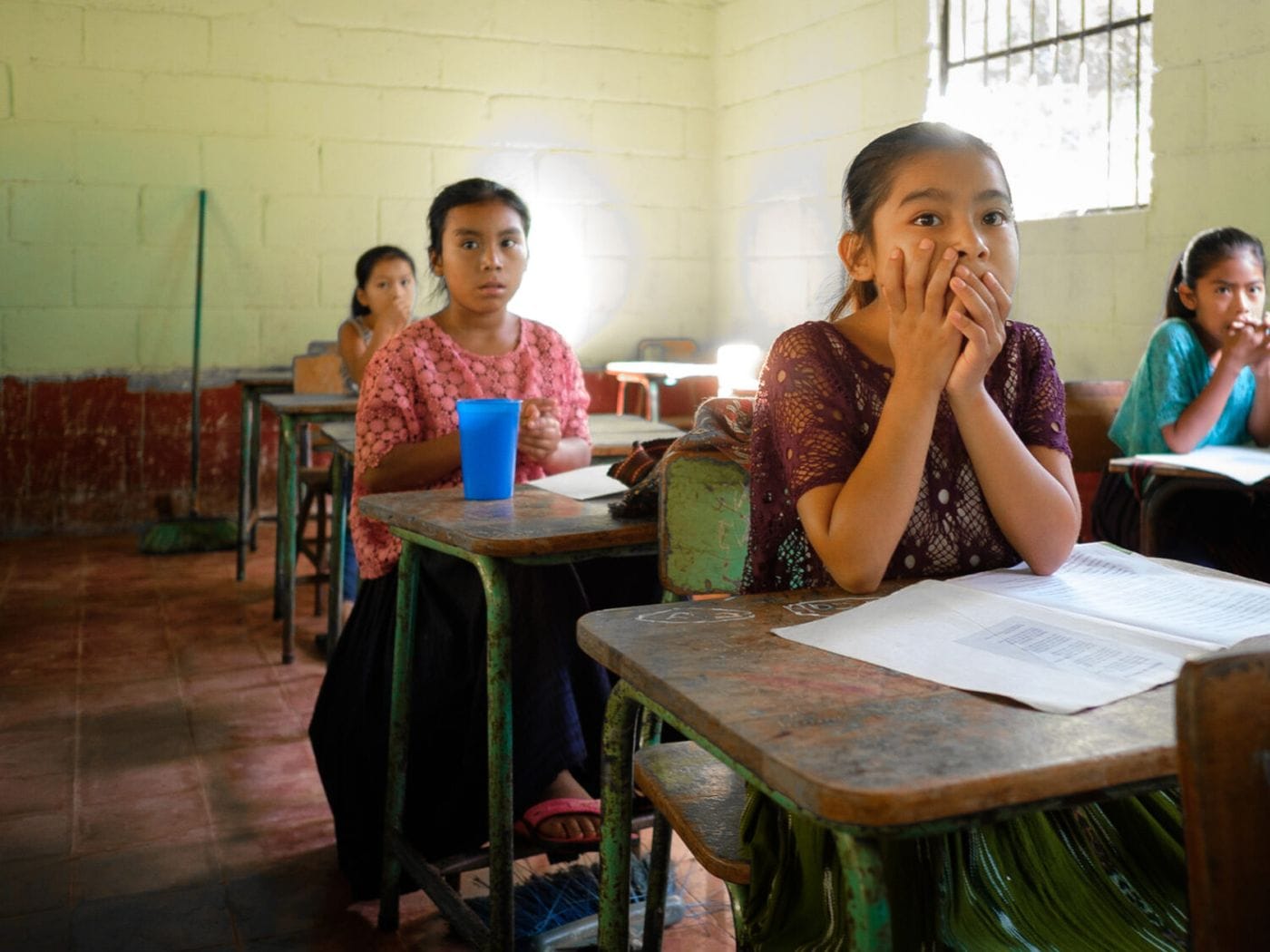 Four young girls sit at desks in a simple classroom, with the girl in the foreground holding her hands to her mouth while looking towards the camera.