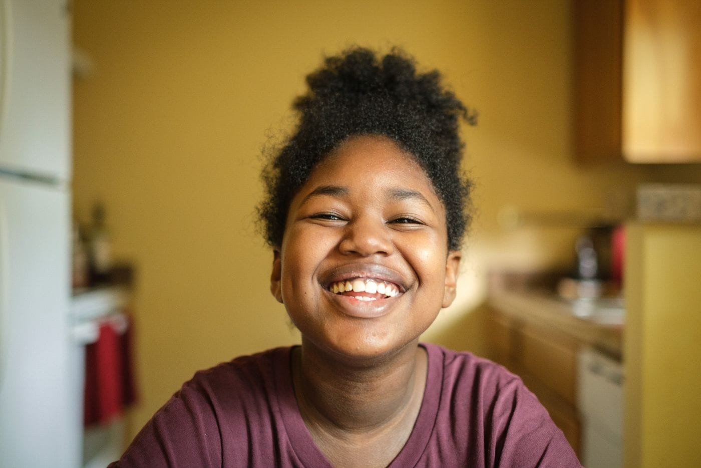 A young person with dark, curly hair pulled into a high bun smiles broadly at the camera while wearing a maroon shirt in a softly blurred kitchen setting.