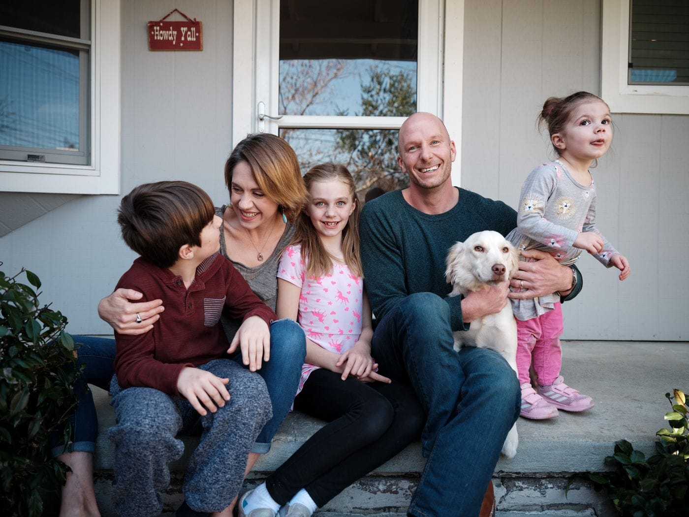 A family of five, including a mother, father, two daughters, a son, and a light-colored dog, sits together on a porch in front of a gray house with a "Howdy Y'all" sign.