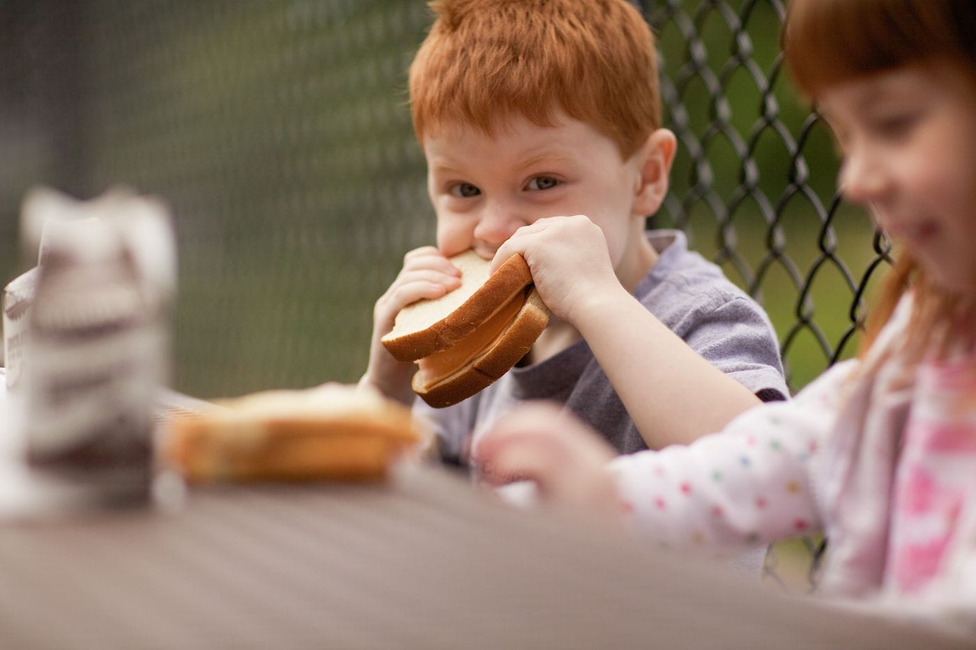 A young boy with red hair eats a sandwich at a picnic table while a blurry girl with red hair sits to his right.