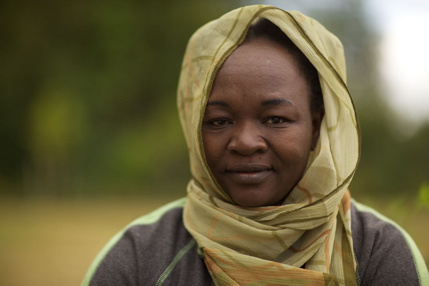 A woman with dark skin wears a light green and tan patterned headscarf and a grey long-sleeved shirt with green accents, looking directly at the camera against a blurred outdoor background.