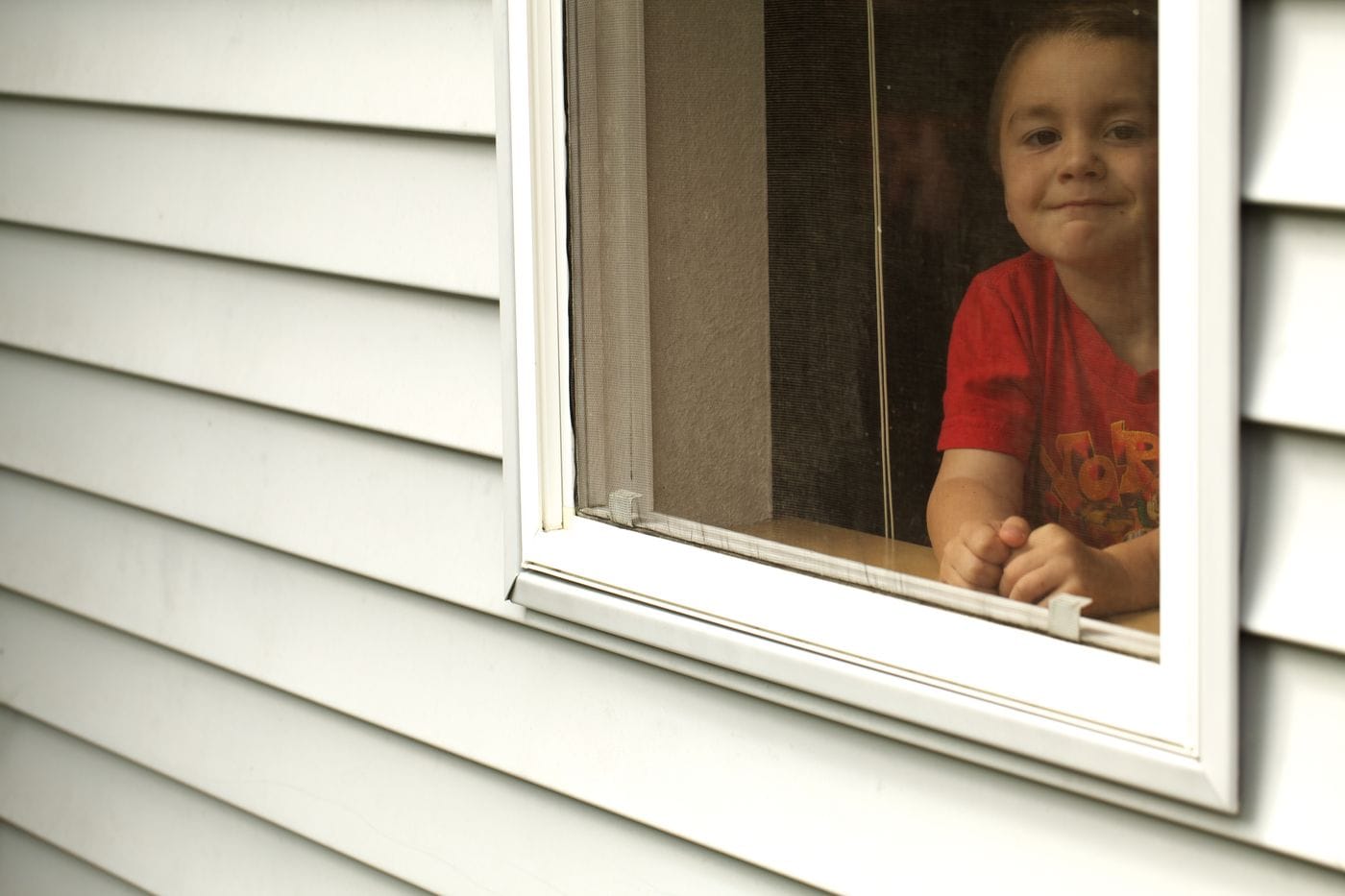 A young boy in a red graphic t-shirt peeks out through a window screen from inside a house with gray vinyl siding.