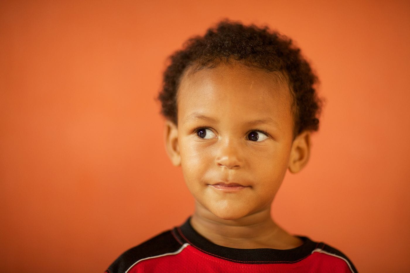 A young child with short curly hair and a curious expression looks to the side against a solid orange background.