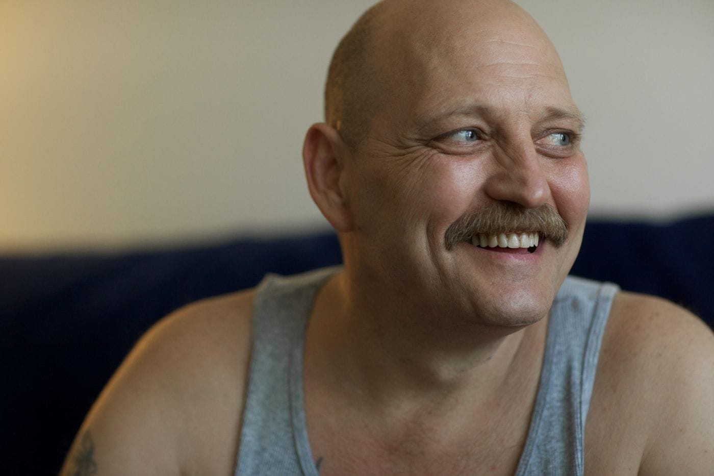 A bald, middle-aged man with a mustache smiles while looking off to the side, wearing a grey ribbed tank top against a simple background.