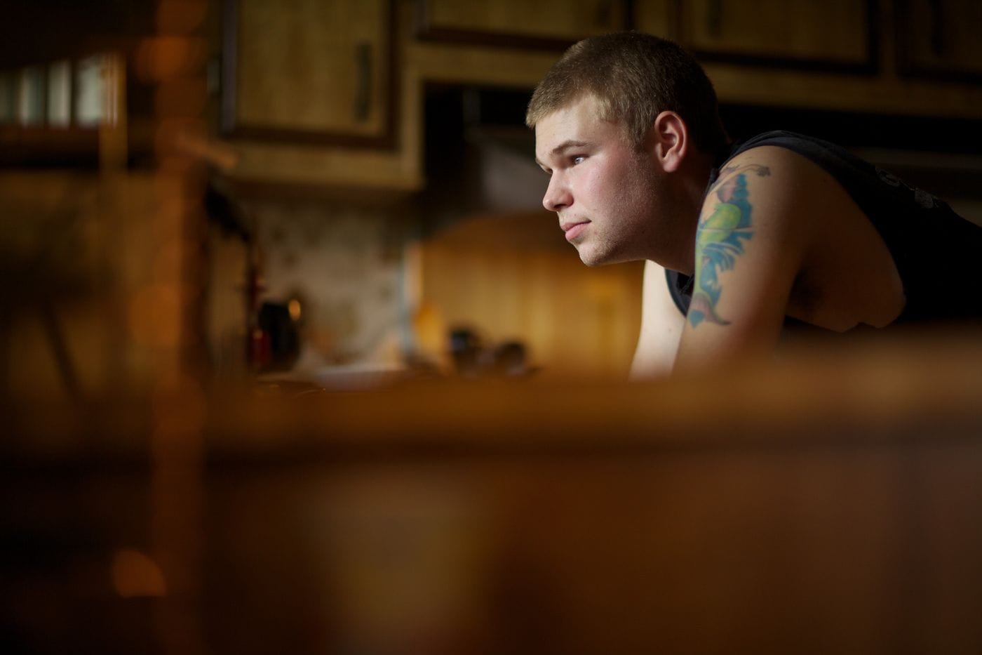 A side profile of a young man with a tattoo on his upper arm looks toward something off-camera while leaning against a counter in a dimly lit, rustic kitchen.