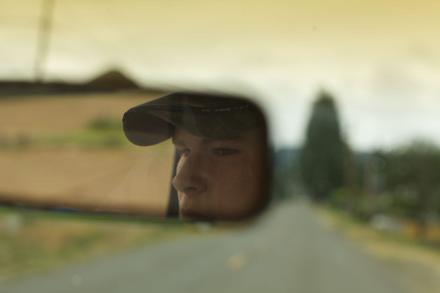 A reflection of a young man wearing a baseball cap is captured in a rearview mirror, against a blurry backdrop of a road and landscape.