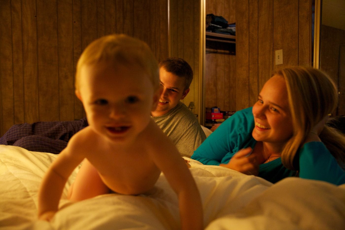 A young blonde toddler crawls on a white bed in the foreground while a man and a smiling woman watch from behind, with wood-paneled walls and an open closet in the background.