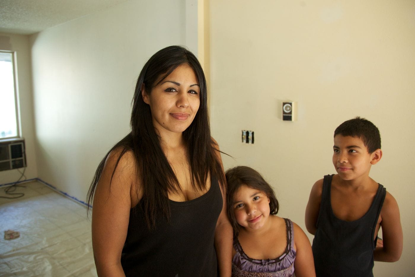 A woman with long dark hair stands indoors next to her young daughter, who leans against her, and her young son, who stands to their right.